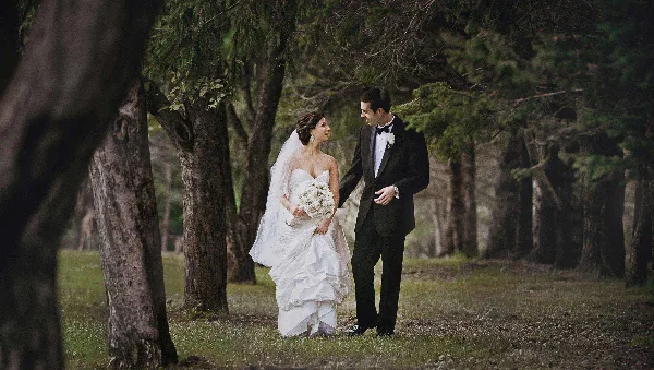 A bride in a white gown and a groom in a tuxedo standing together in a wooded area.