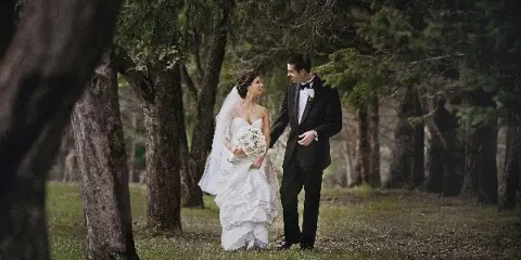 A bride in a white gown and a groom in a tuxedo standing together in a wooded area.