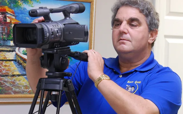 A man in a blue polo shirt with a 'Sam Gold' logo operating a professional Sony video camera mounted on a tripod.