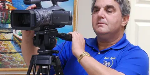 A man in a blue polo shirt with a 'Sam Gold' logo operating a professional Sony video camera mounted on a tripod.