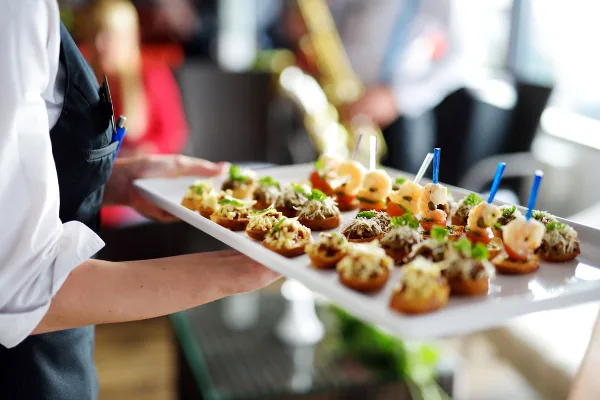 A catering server in a black apron holding a white platter of shrimp and vegetable bruschetta appetizers.
