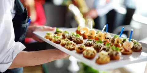 A catering server in a black apron holding a white platter of shrimp and vegetable bruschetta appetizers.