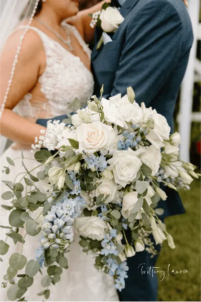 A close-up of a bride holding a cascading wedding bouquet with white roses, blue delphiniums, and eucalyptus.