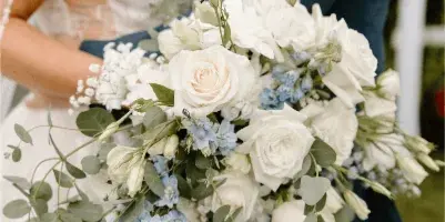 A close-up of a bride holding a cascading wedding bouquet with white roses, blue delphiniums, and eucalyptus.