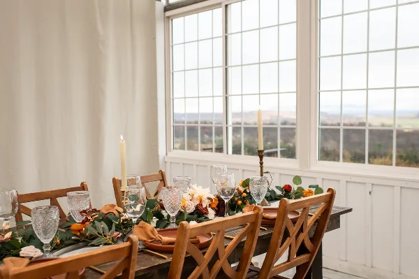 A rustic wooden table set with a floral and greenery garland, glassware, and candles in a bright room with large windows.