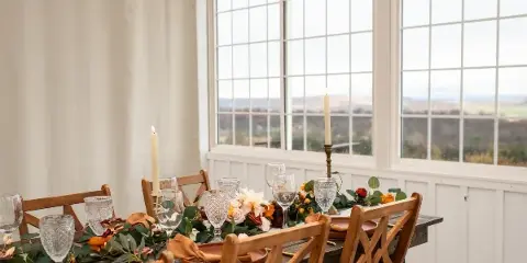 A rustic wooden table set with a floral and greenery garland, glassware, and candles in a bright room with large windows.