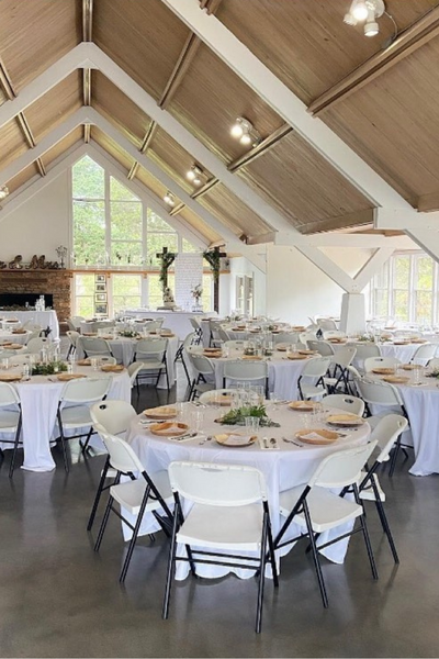 Interior view of a large wedding reception hall with white tables and chairs set up under a high wooden ceiling.