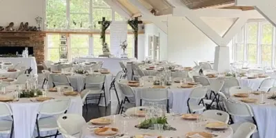 Interior view of a large wedding reception hall with white tables and chairs set up under a high wooden ceiling.