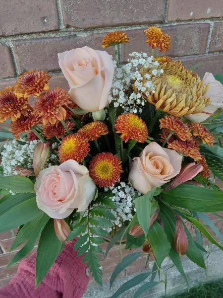 A hand holding a floral arrangement with peach roses, orange chrysanthemums, and white baby's breath against a brick wall.