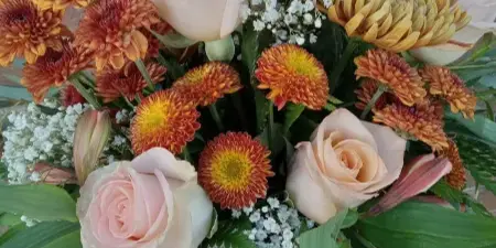 A hand holding a floral arrangement with peach roses, orange chrysanthemums, and white baby's breath against a brick wall.