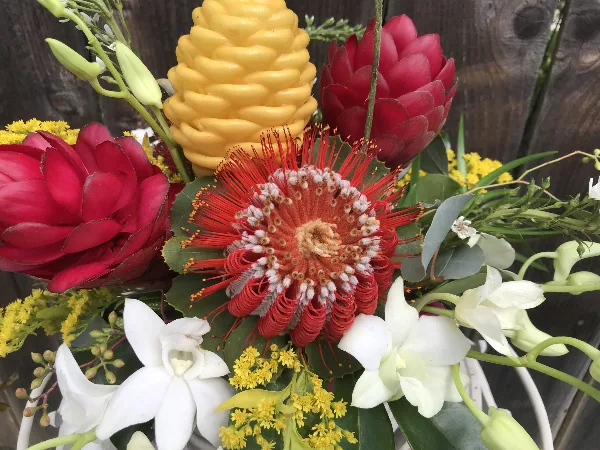 A close-up of a tropical floral arrangement featuring red and yellow proteas, white orchids, and yellow filler flowers.