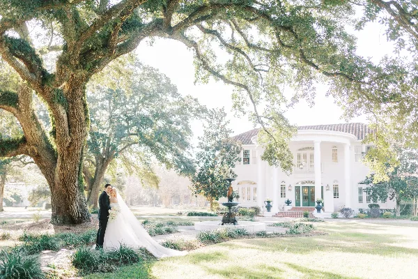 A bride and groom standing together in a garden with a large white mansion in the background.