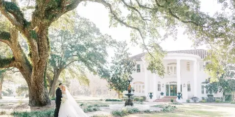 A bride and groom standing together in a garden with a large white mansion in the background.
