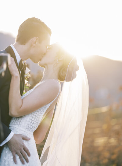 A bride and groom kissing outdoors at sunset with a mountainous background.