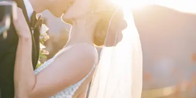 A bride and groom kissing outdoors at sunset with a mountainous background.
