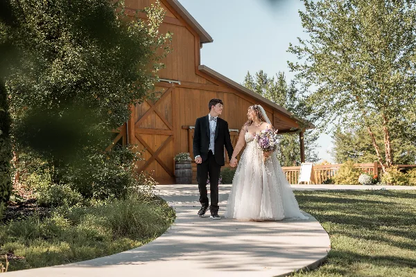 A bride and groom holding hands and walking on a paved path near a wooden barn structure.