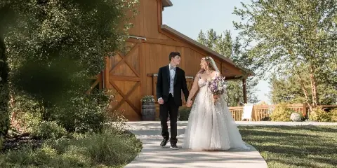 A bride and groom holding hands and walking on a paved path near a wooden barn structure.