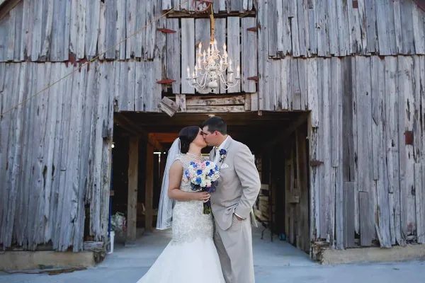 A bride and groom kissing in front of a rustic wooden barn with a chandelier hanging above them.