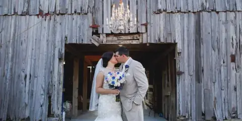 A bride and groom kissing in front of a rustic wooden barn with a chandelier hanging above them.