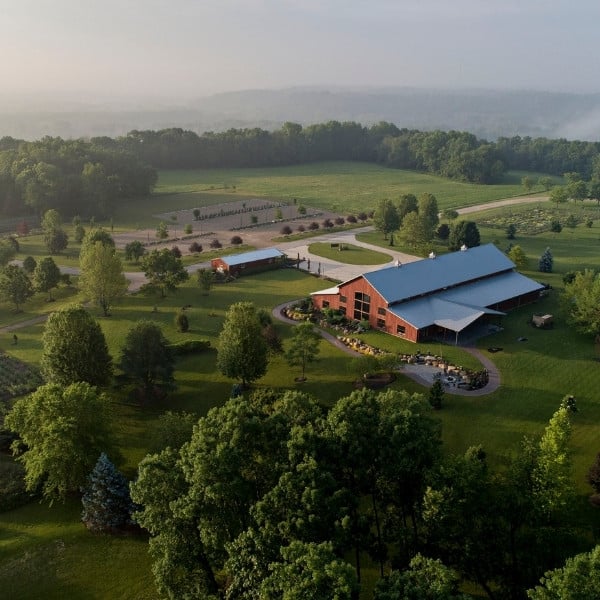 Aerial view of a red barn with a metal roof surrounded by green fields, trees, and garden pathways.