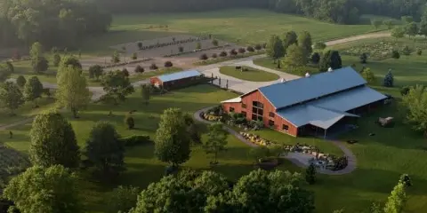 Aerial view of a red barn with a metal roof surrounded by green fields, trees, and garden pathways.