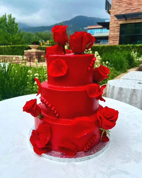 Three-tier red wedding cake decorated with red roses, photographed outdoors with mountains behind it.
