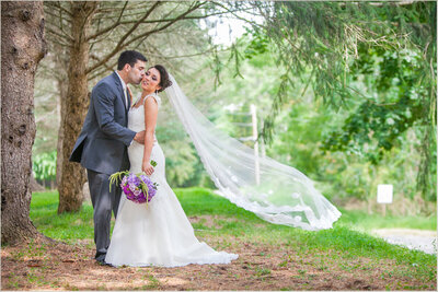 A bride and groom embracing outdoors in a wooded area, with the bride's veil blowing in the wind.