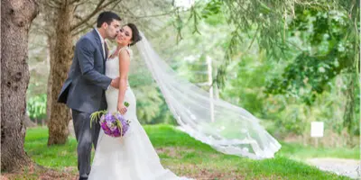 A bride and groom embracing outdoors in a wooded area, with the bride's veil blowing in the wind.