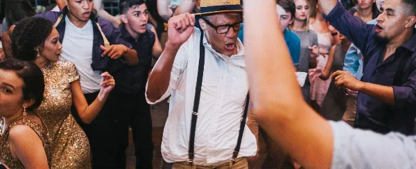 A group of people dancing and celebrating at a lively event, with a man in a straw hat and suspenders in the center.