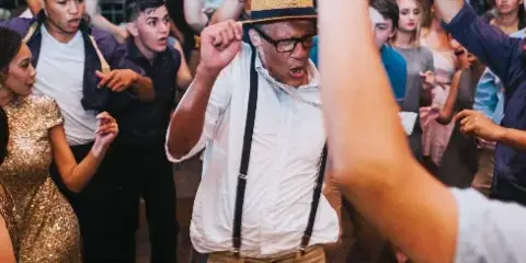 A group of people dancing and celebrating at a lively event, with a man in a straw hat and suspenders in the center.