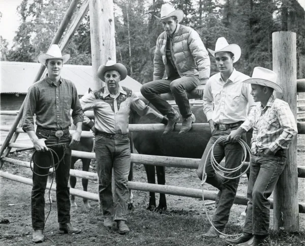 A group of five men in cowboy hats and western attire posing with a horse and lasso in a wooden corral.
