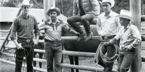 A group of five men in cowboy hats and western attire posing with a horse and lasso in a wooden corral.