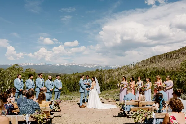 Outdoor wedding ceremony in the Rocky Mountains with a bride and groom exchanging vows surrounded by wedding party and guests.