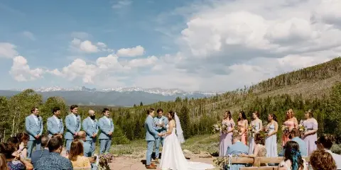 Outdoor wedding ceremony in the Rocky Mountains with a bride and groom exchanging vows surrounded by wedding party and guests.