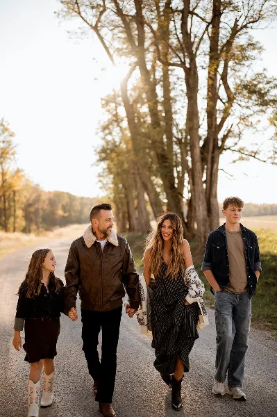 A family of four walking on a gravel road in a rural setting, with the woman in the center wearing a striped dress and holding a patterned shawl.