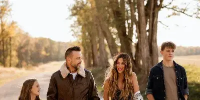 A family of four walking on a gravel road in a rural setting, with the woman in the center wearing a striped dress and holding a patterned shawl.