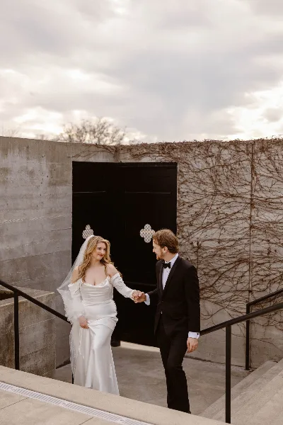 A bride in a white gown and a groom in a black tuxedo holding hands on a concrete staircase.