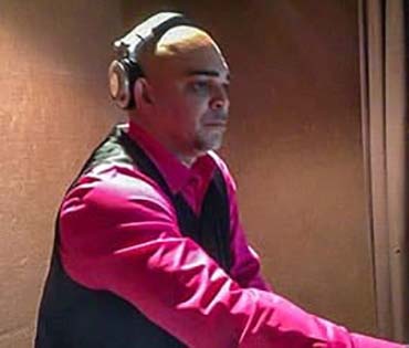 A professional DJ wearing headphones and a pink shirt, seated at a mixing console.