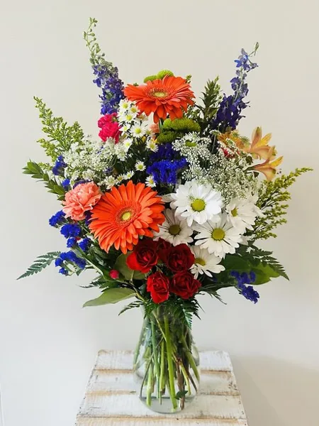 A colorful floral arrangement featuring orange gerbera daisies, red roses, white daisies, and blue delphiniums in a clear glass vase.
