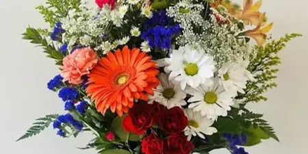 A colorful floral arrangement featuring orange gerbera daisies, red roses, white daisies, and blue delphiniums in a clear glass vase.