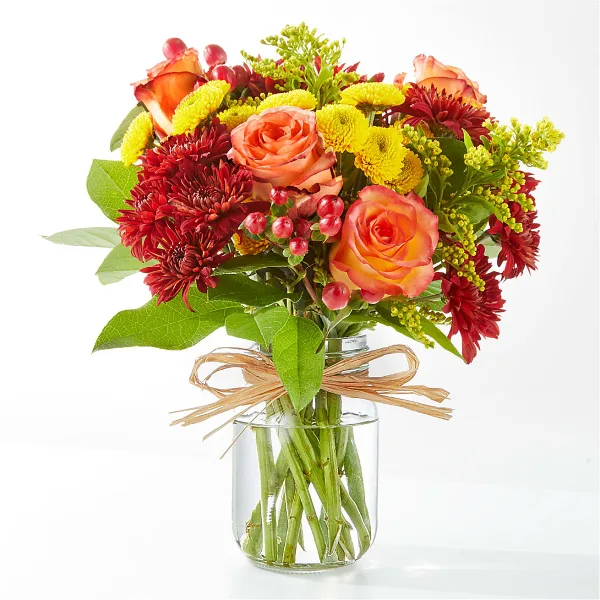 A colorful floral arrangement featuring orange roses, red chrysanthemums, and yellow filler flowers in a clear glass vase.