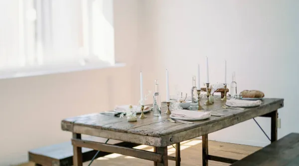 A rustic wooden table set with white candles, glassware, and folded napkins in a bright indoor venue space.