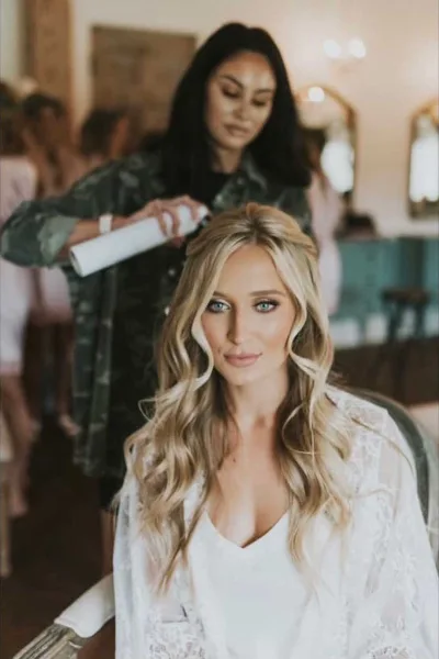Bridal stylist working on a brides hair while the bride sits in a white robe.