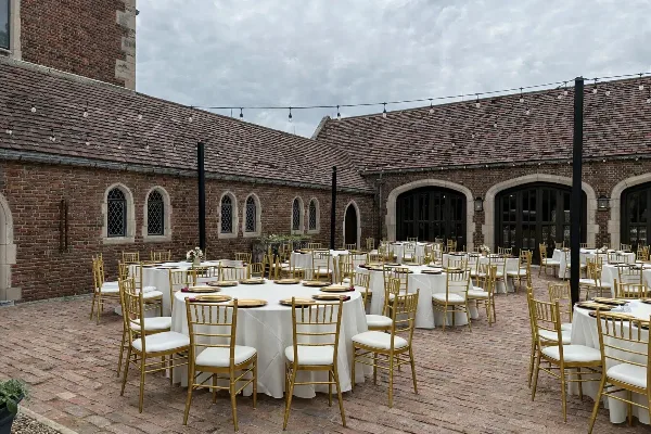 Outdoor wedding reception setup featuring gold Chiavari chairs, white table linens, and charger plates in a brick courtyard.