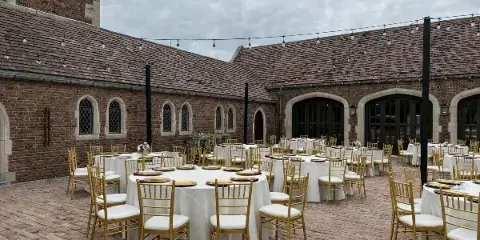 Outdoor wedding reception setup featuring gold Chiavari chairs, white table linens, and charger plates in a brick courtyard.