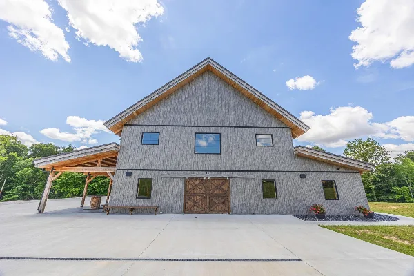 Exterior view of a large rustic event barn with grey siding, a metal roof, and a covered porch area under a blue sky.