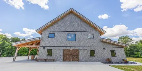 Exterior view of a large rustic event barn with grey siding, a metal roof, and a covered porch area under a blue sky.