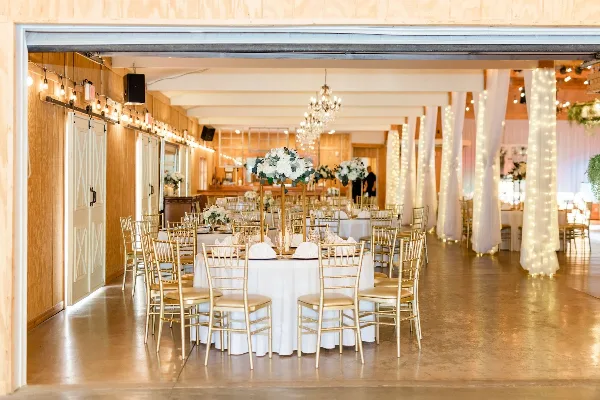 Interior of a rustic banquet hall set up for a wedding reception with round tables, gold chairs, white floral centerpieces, and string lights.