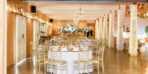 Interior of a rustic banquet hall set up for a wedding reception with round tables, gold chairs, white floral centerpieces, and string lights.