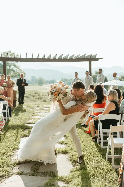 A bride and groom share a kiss during an outdoor wedding ceremony with an officiant and wedding party present, set against a backdrop of mountains.
